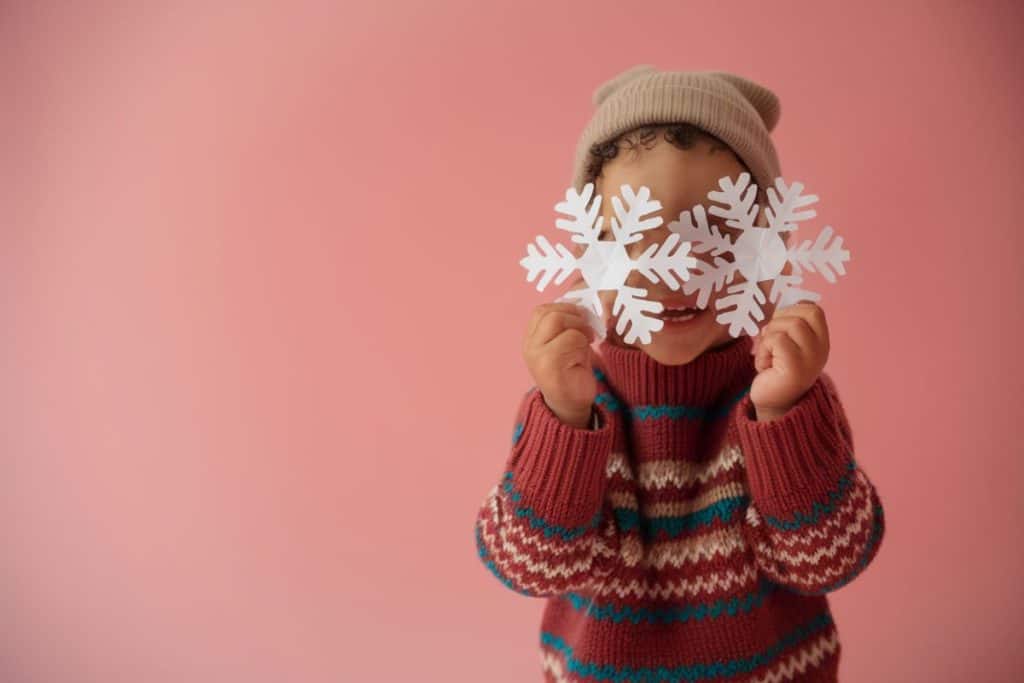 Et smilende barn med stribet sweater og beige hue holder papir-snefnug op foran ansigtet.